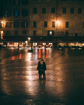 A couple embraces on a dimly lit Italian piazza at night with reflections of warm lights.