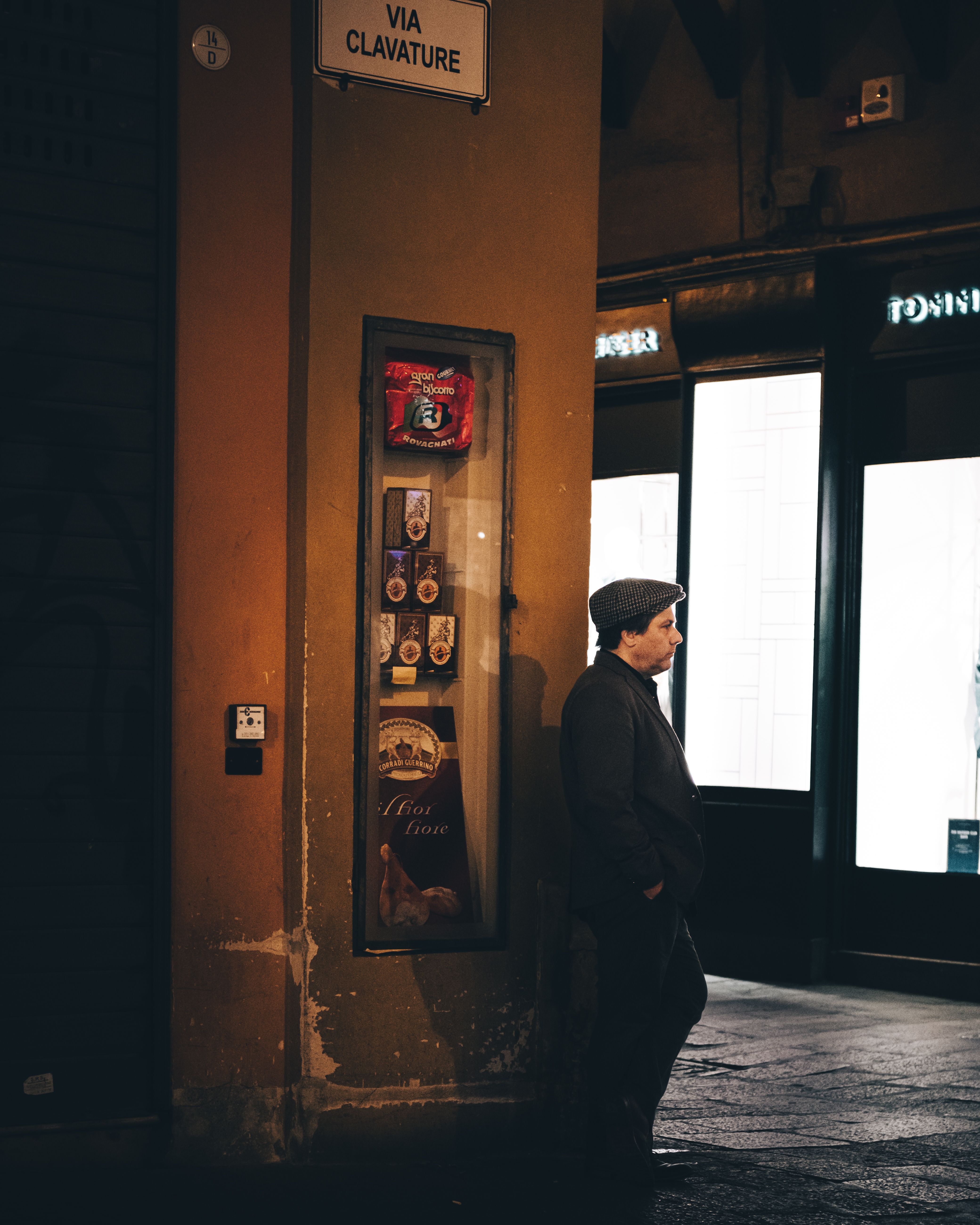 A man in a cap leans against a wall at dusk in a city street near a Via Clavature sign.