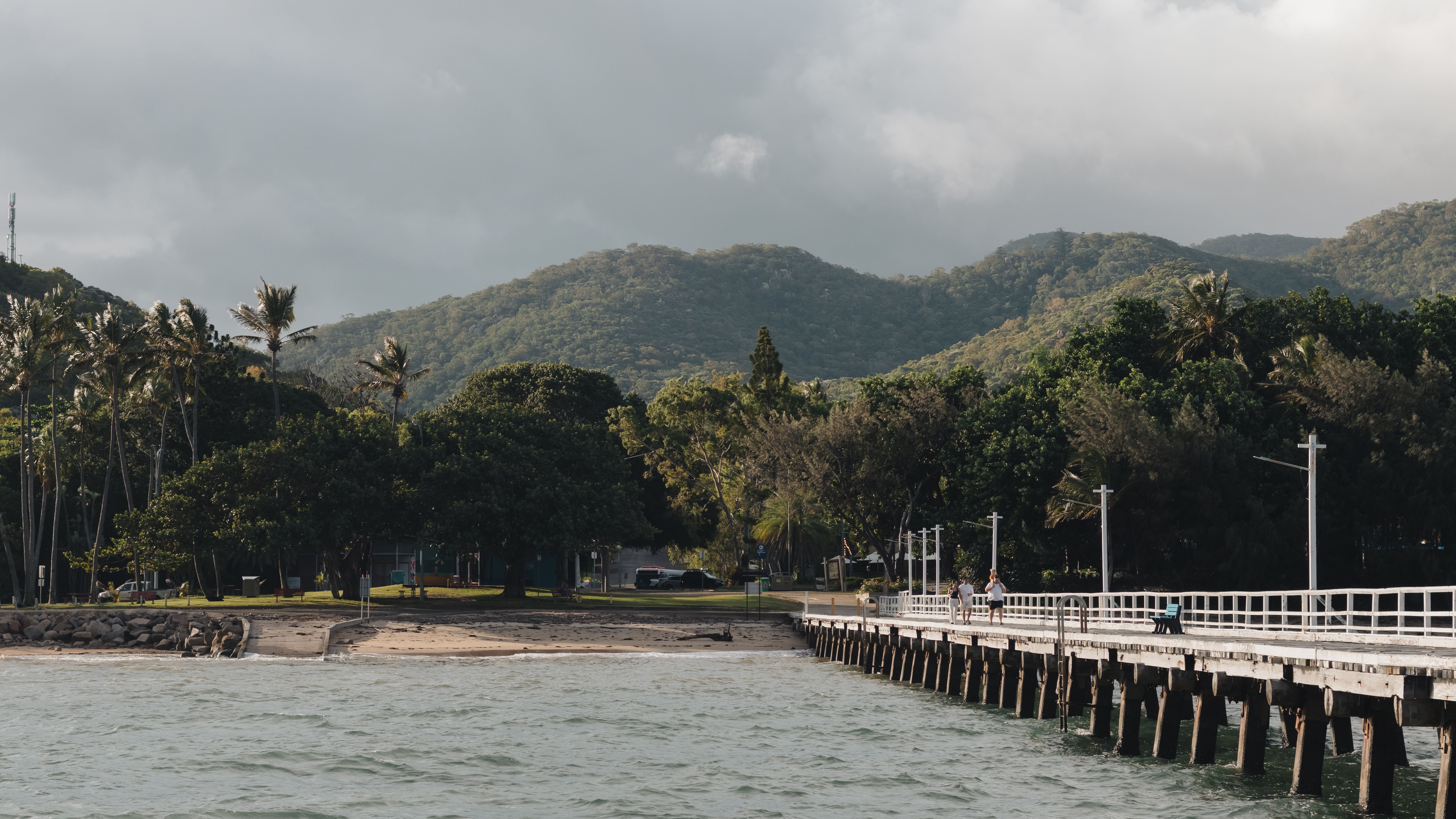 Scenic view of a coastal pier and lush mountains under a cloudy sky in Queensland, Australia.