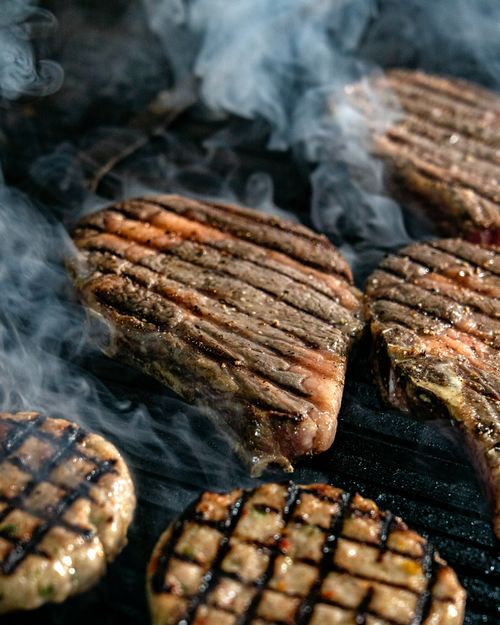 Free Close-up of sizzling steaks and burgers cooking on an outdoor grill with visible smoke. Stock Photo