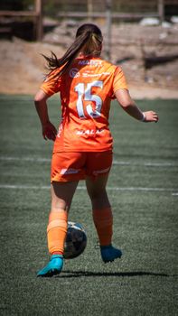Action shot of a female soccer player wearing orange, dribbling the ball on a sunny day.
