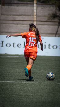 Female soccer player in action on outdoor field with orange uniform, captured from behind.