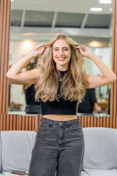 A cheerful woman with long blonde hair poses confidently in a salon setting.