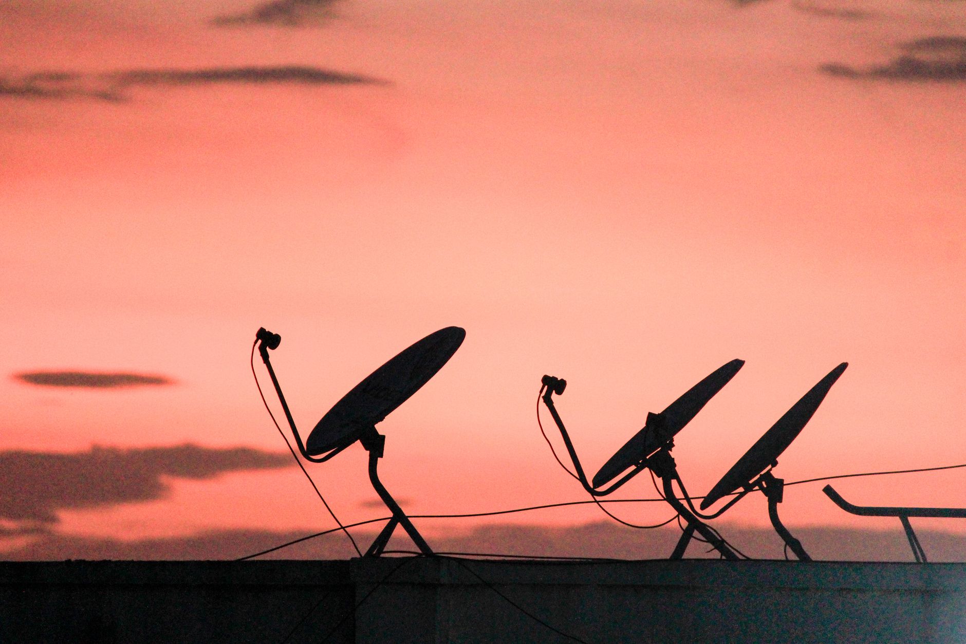 A row of satellite dishes silhouetted against a vibrant orange sunset sky on a rooftop.