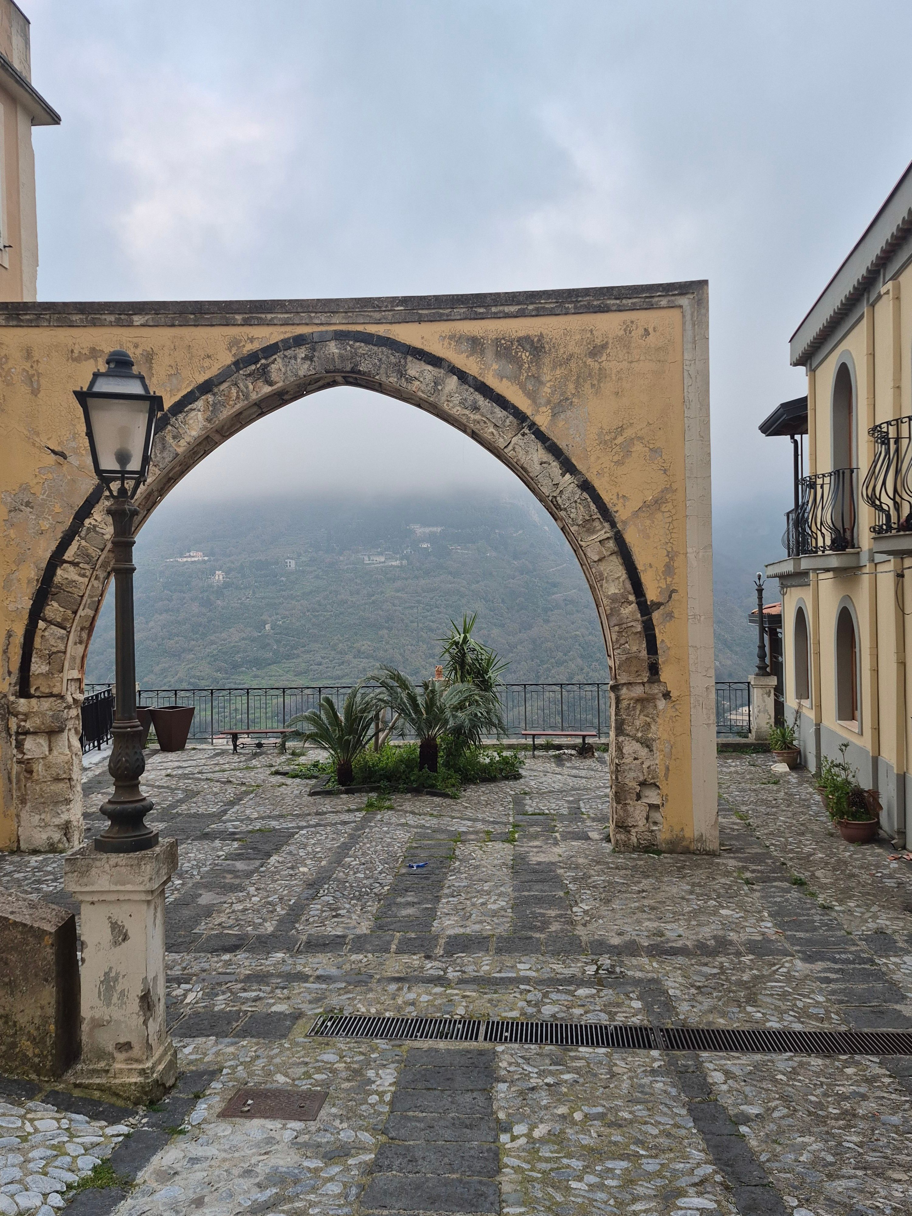Fotomural Una vista pintoresca a través de un arco de piedra en Castroreale, Sicilia, Italia, con paisaje montañoso.