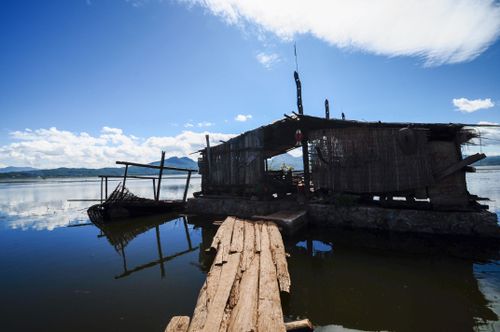 Free A rustic fishing hut on a serene lake, with a wooden dock under a bright blue sky. Stock Photo