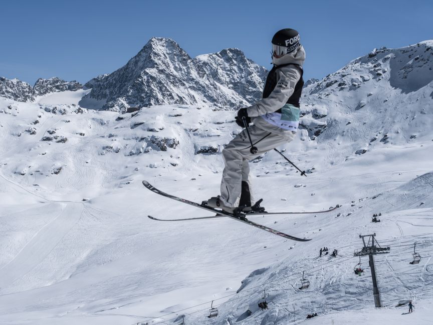 A skier jumps high in the air against a backdrop of snowy mountains and ski lifts in winter.