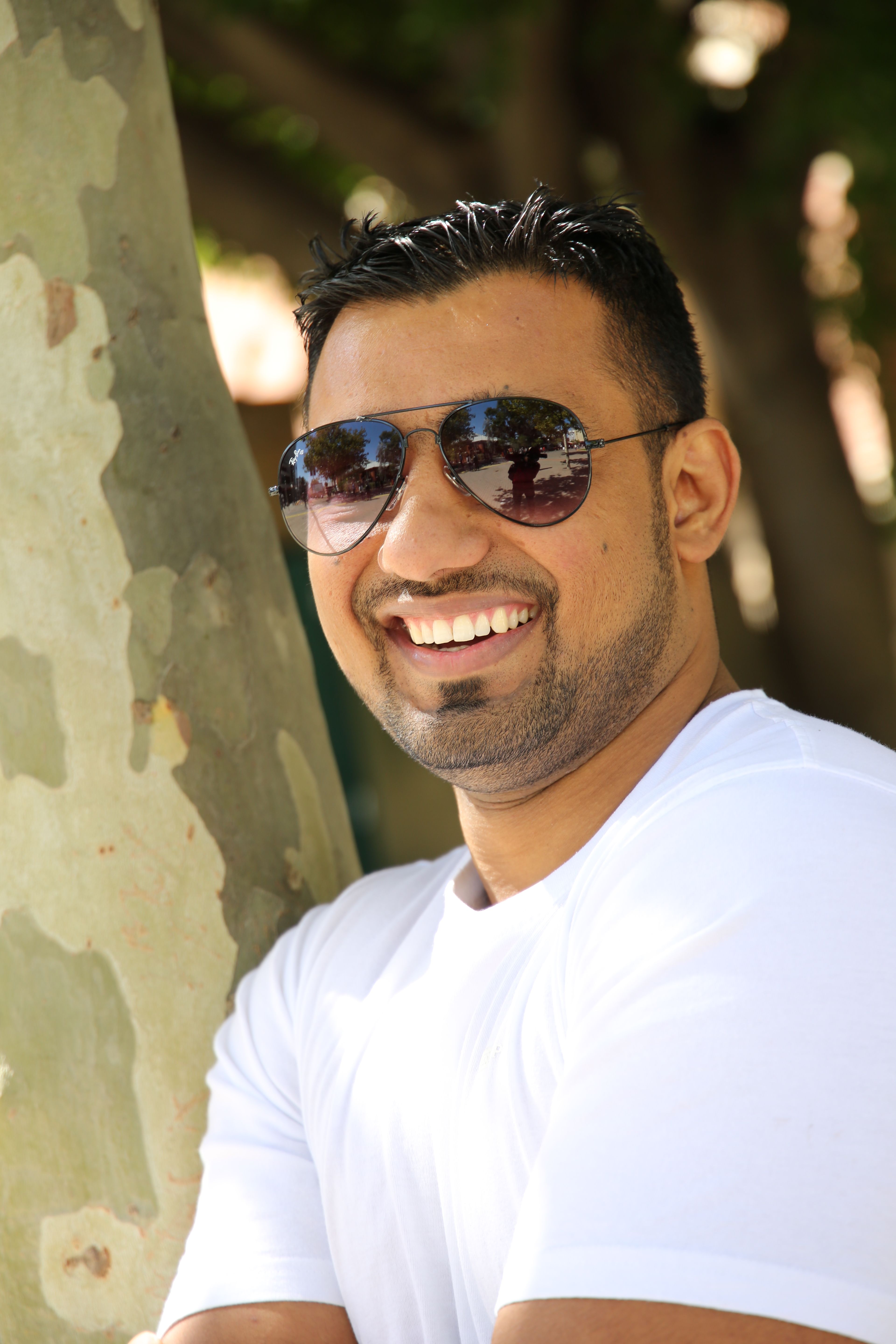 Confident man smiling in sunglasses and white shirt against a tree.