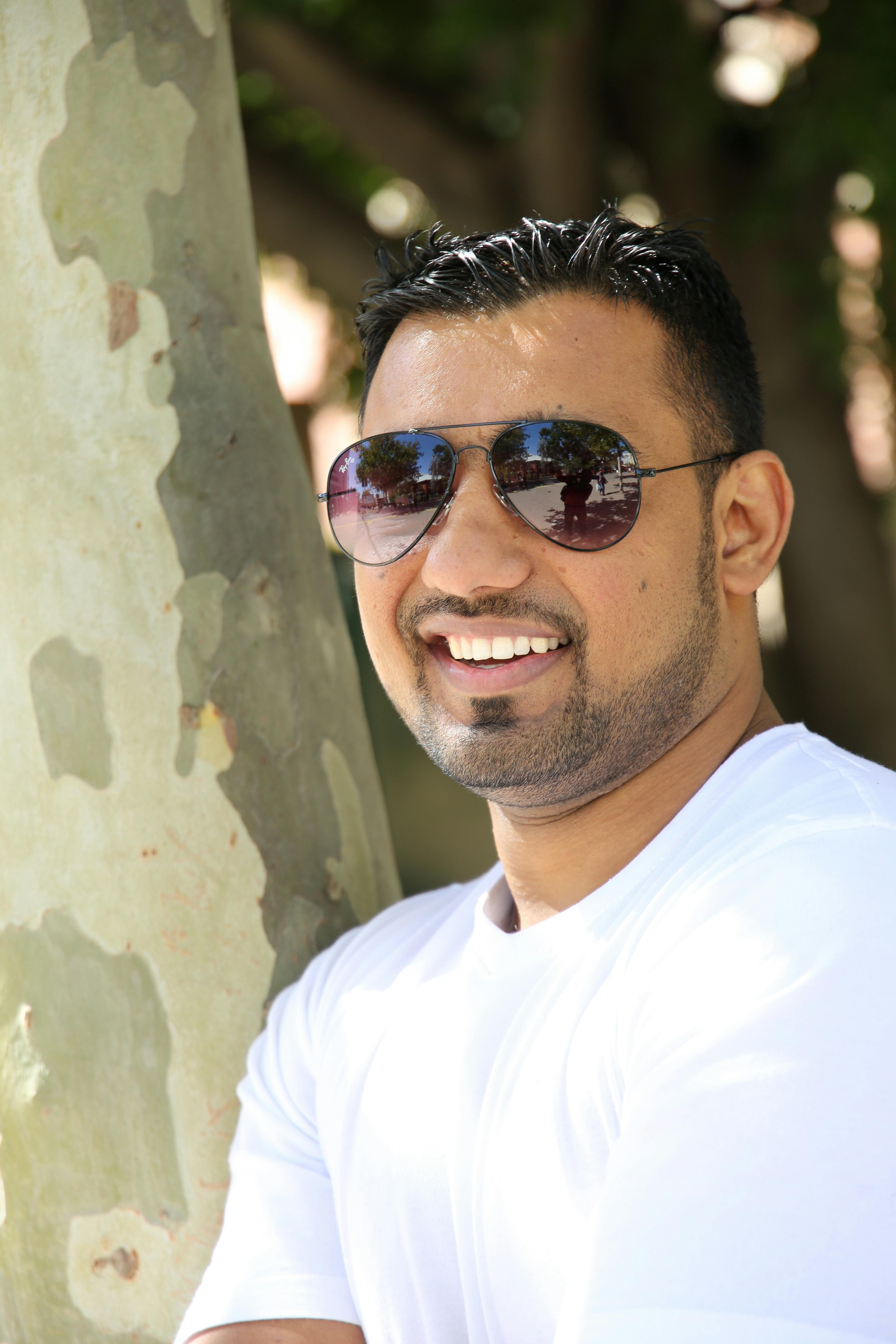 A cheerful man wearing sunglasses and a white shirt, leaning on a tree outdoors.