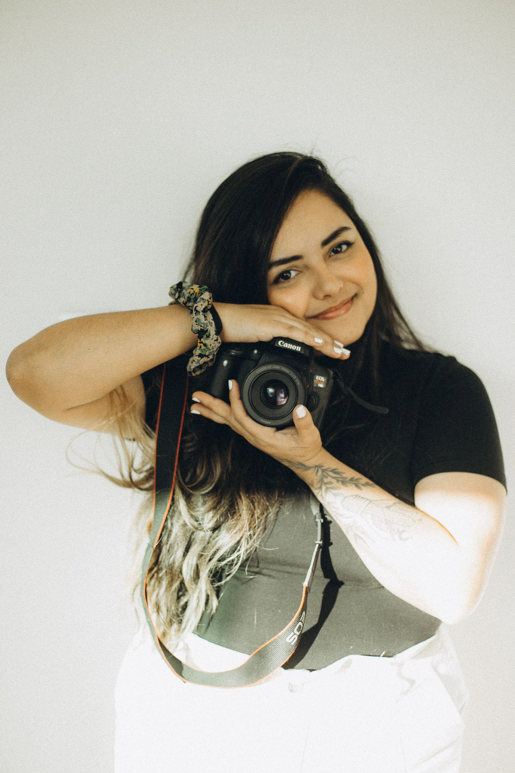 Smiling woman with long hair holding a DSLR camera against a plain background.