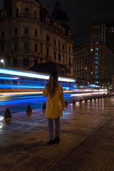 A woman in a yellow coat watches city traffic light trails on a rainy night.
