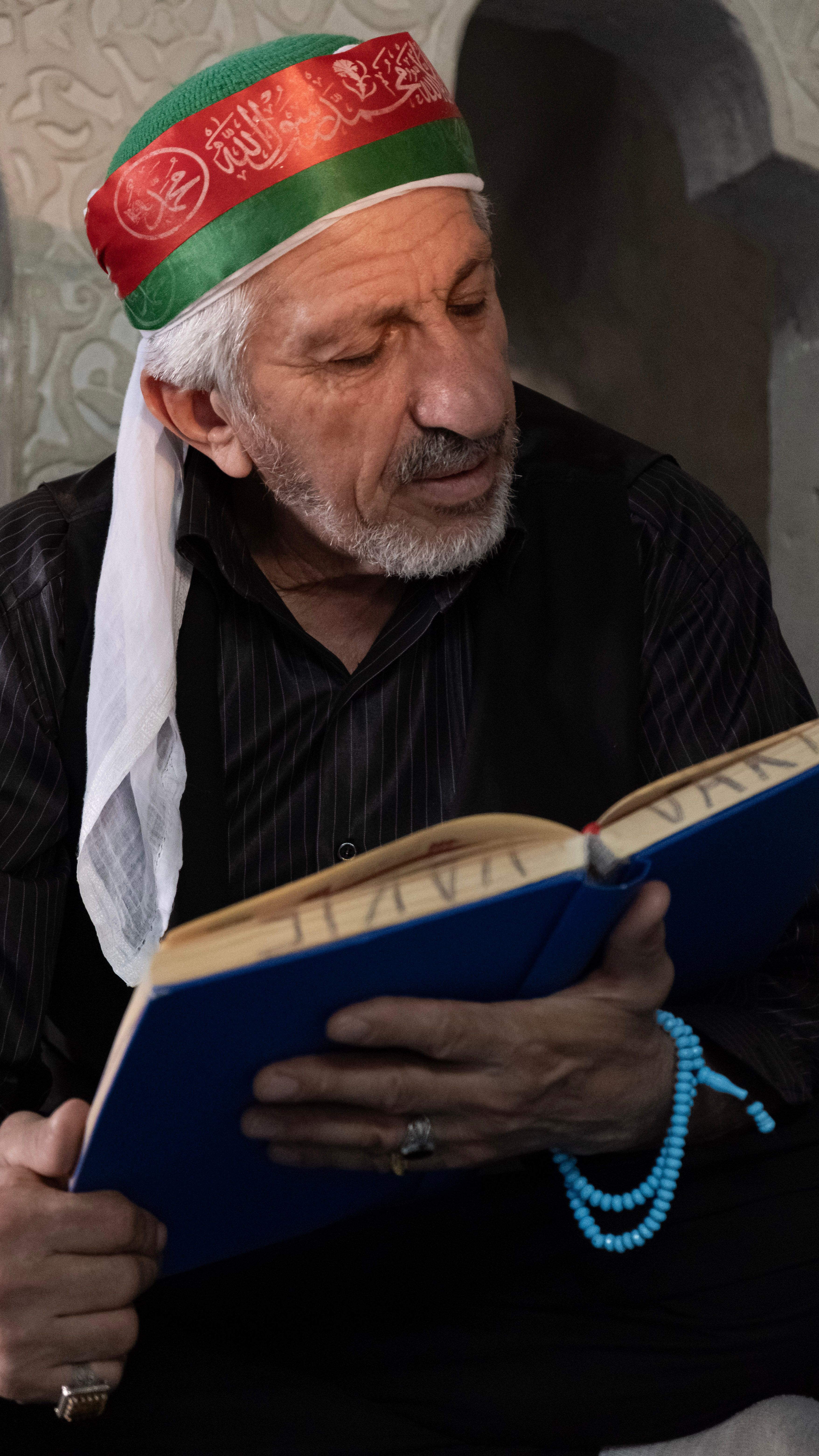 An elderly man wearing a red headscarf reads a book in a dimly lit room in Diyarbakır, Türkiye.