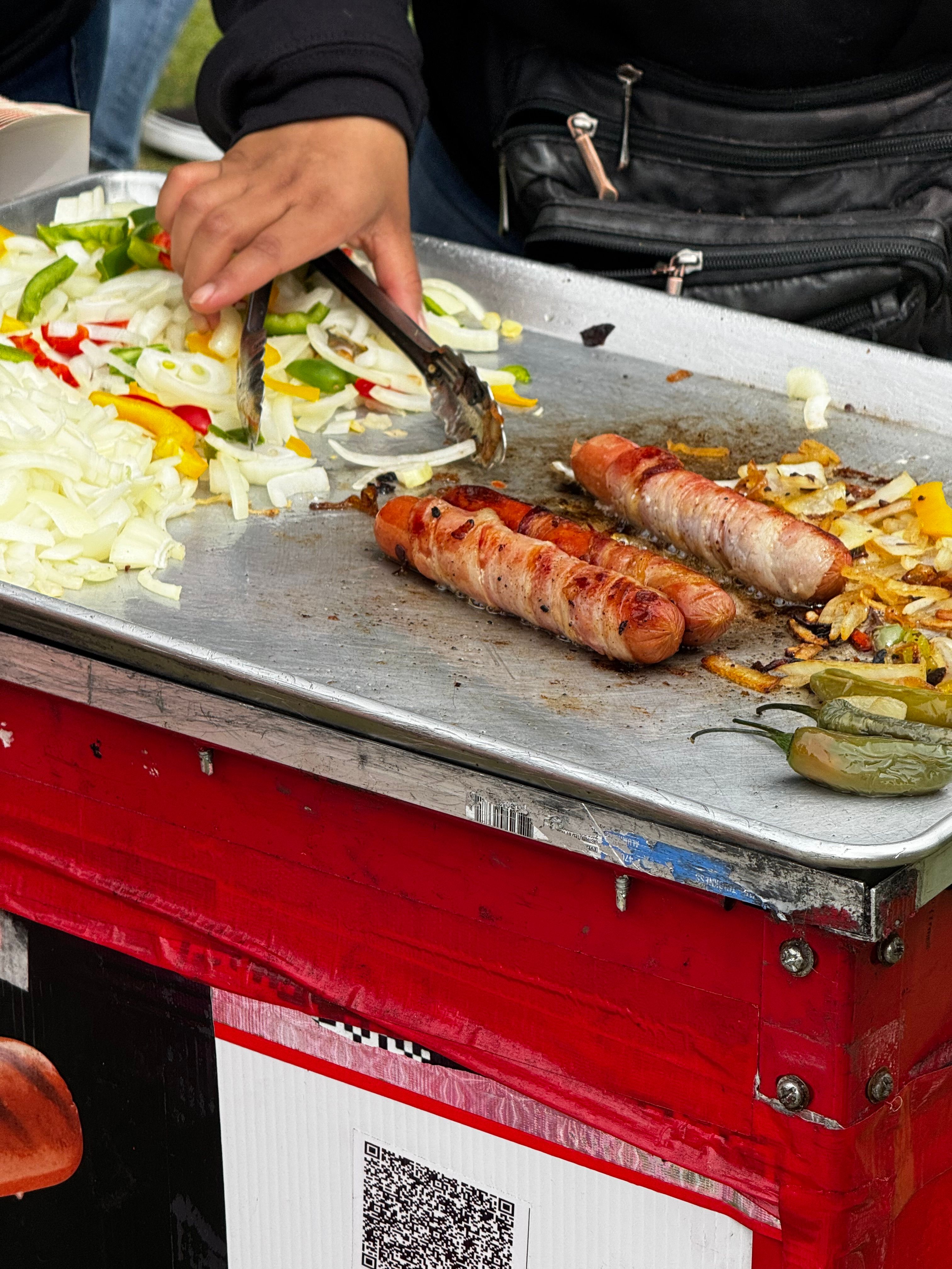 Vibrant street scene of a vendor grilling bacon-wrapped sausages with vegetables.