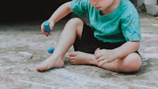 一个年幼的孩子坐在户外地上，用彩色粉笔玩着创意游戏