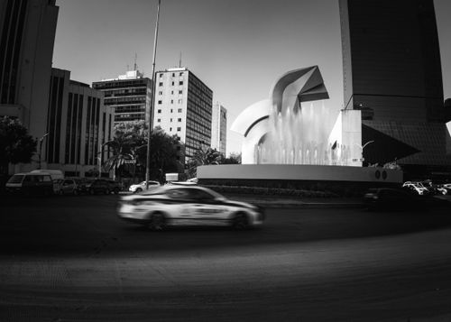 Free Black and white cityscape featuring a modern fountain, motion-blurred car, and tall buildings. Stock Photo
