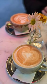 Two cups of heart-shaped latte art with a flower vase on a marble table, outdoors.