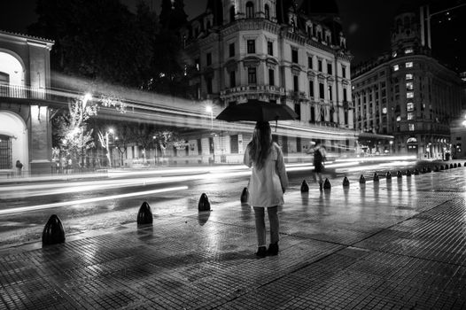 A woman with an umbrella stands on a rainy night street with light trails in Buenos Aires.