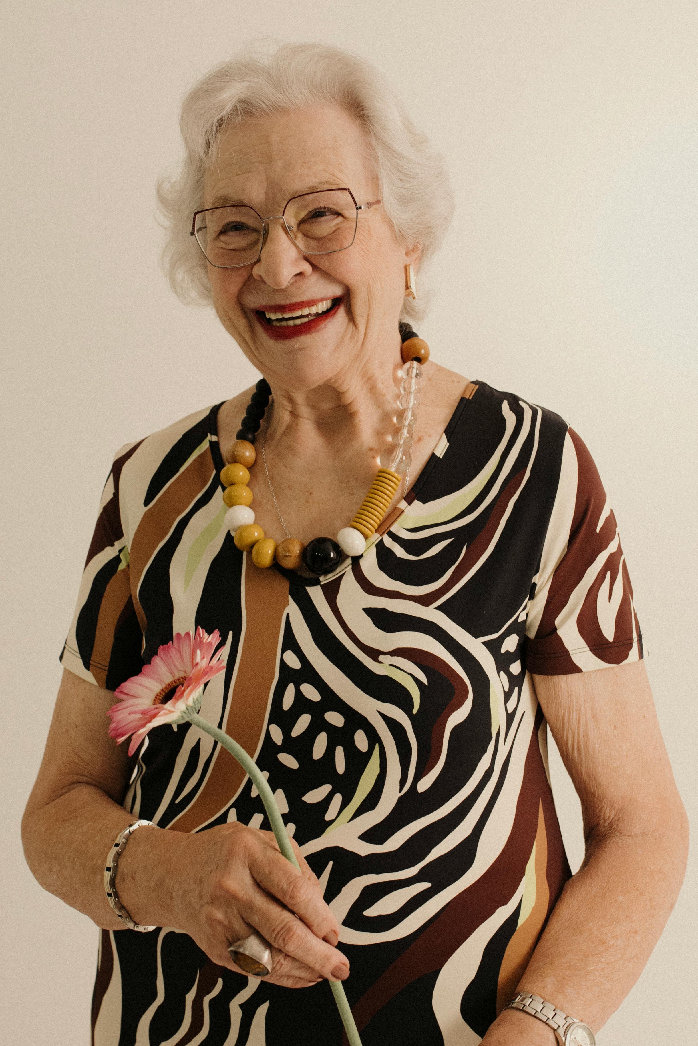 Joyful senior woman in patterned dress holding a pink gerbera flower and smiling warmly.