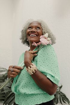A cheerful senior woman with gray hair holding pink flowers while smiling indoors.