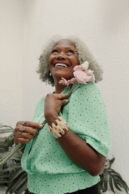 A cheerful senior woman with gray hair holding pink flowers while smiling indoors.