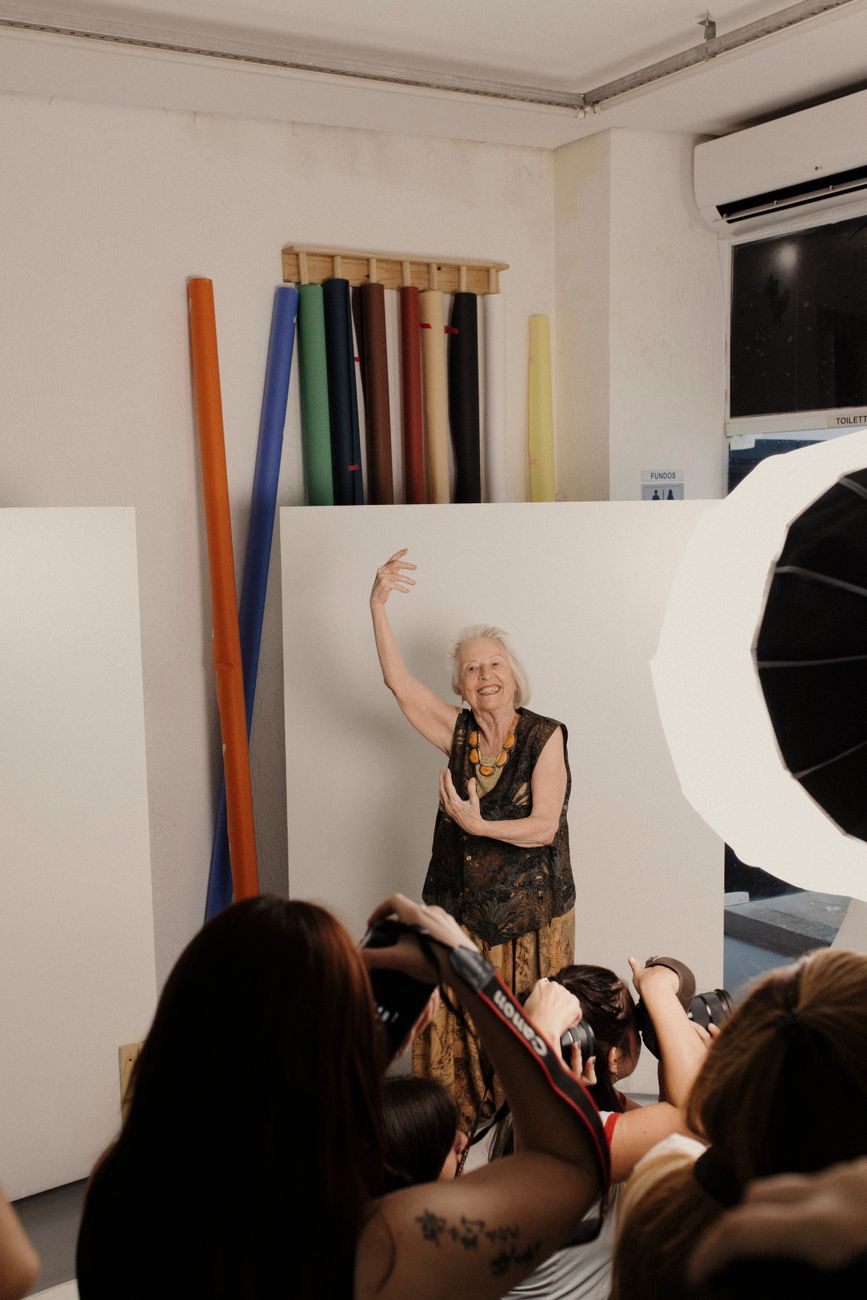 Une femme âgée souriante pose avec élégance devant plusieurs photographes dans un studio, avec des fonds en papier colorés à l'arrière-plan.