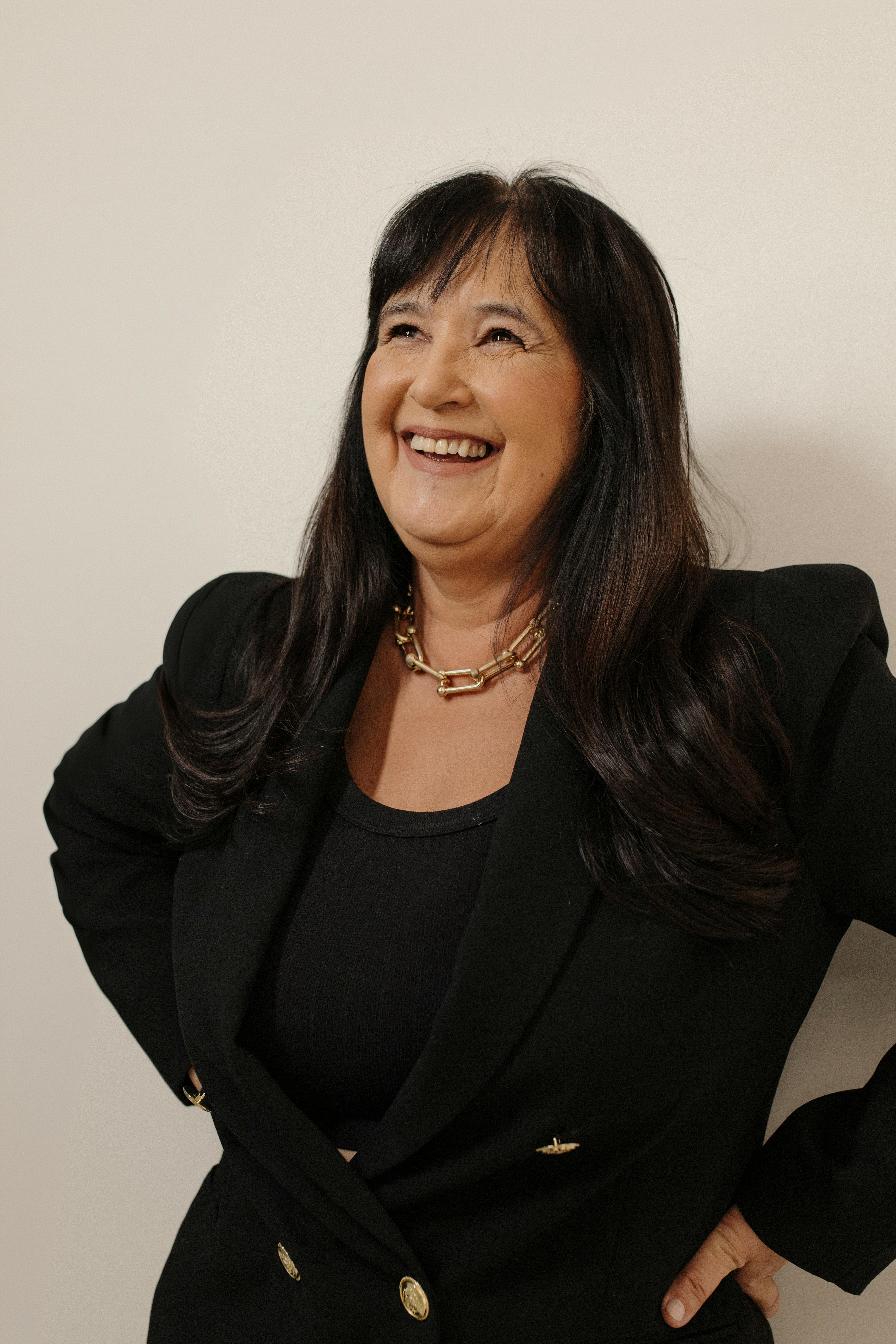 A smiling woman confidently poses in a stylish black blazer against a neutral background.