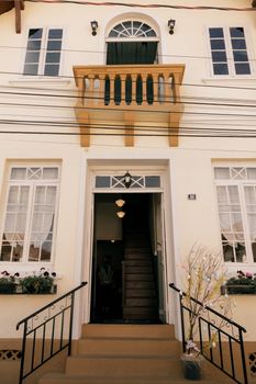 Charming colonial-style house entrance with balcony, featuring decorative windows and lush planters.