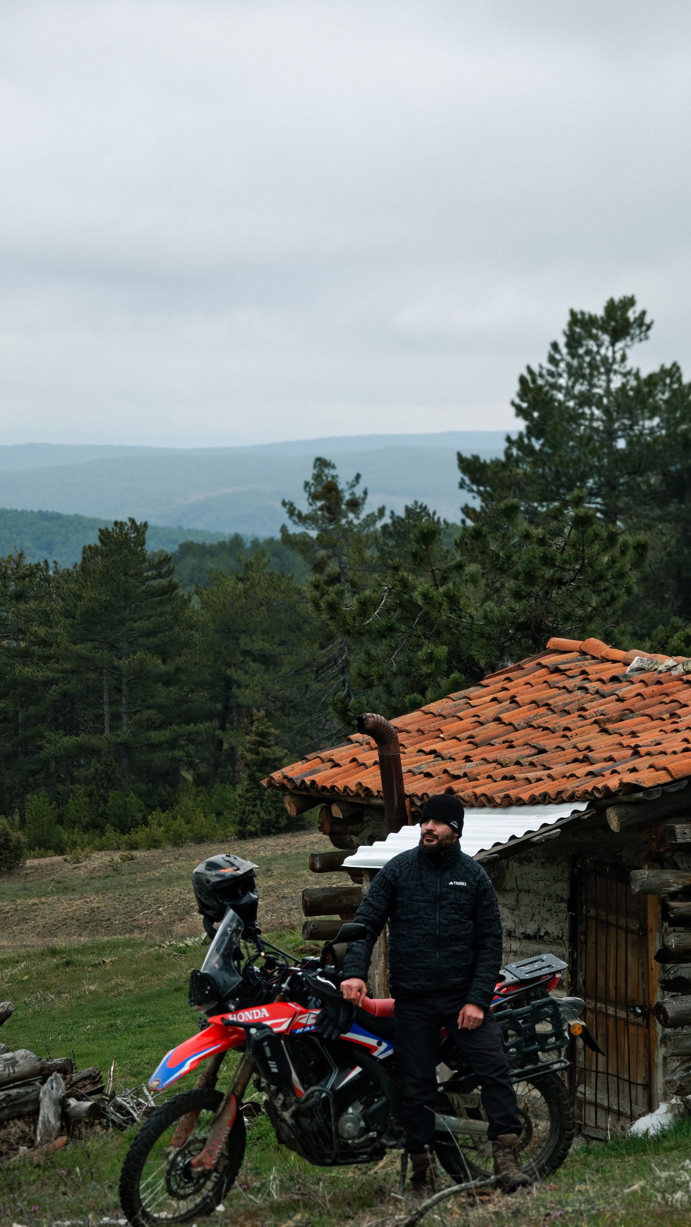 Man with motorcycle beside a wooden hut in a mountainous forest area.
