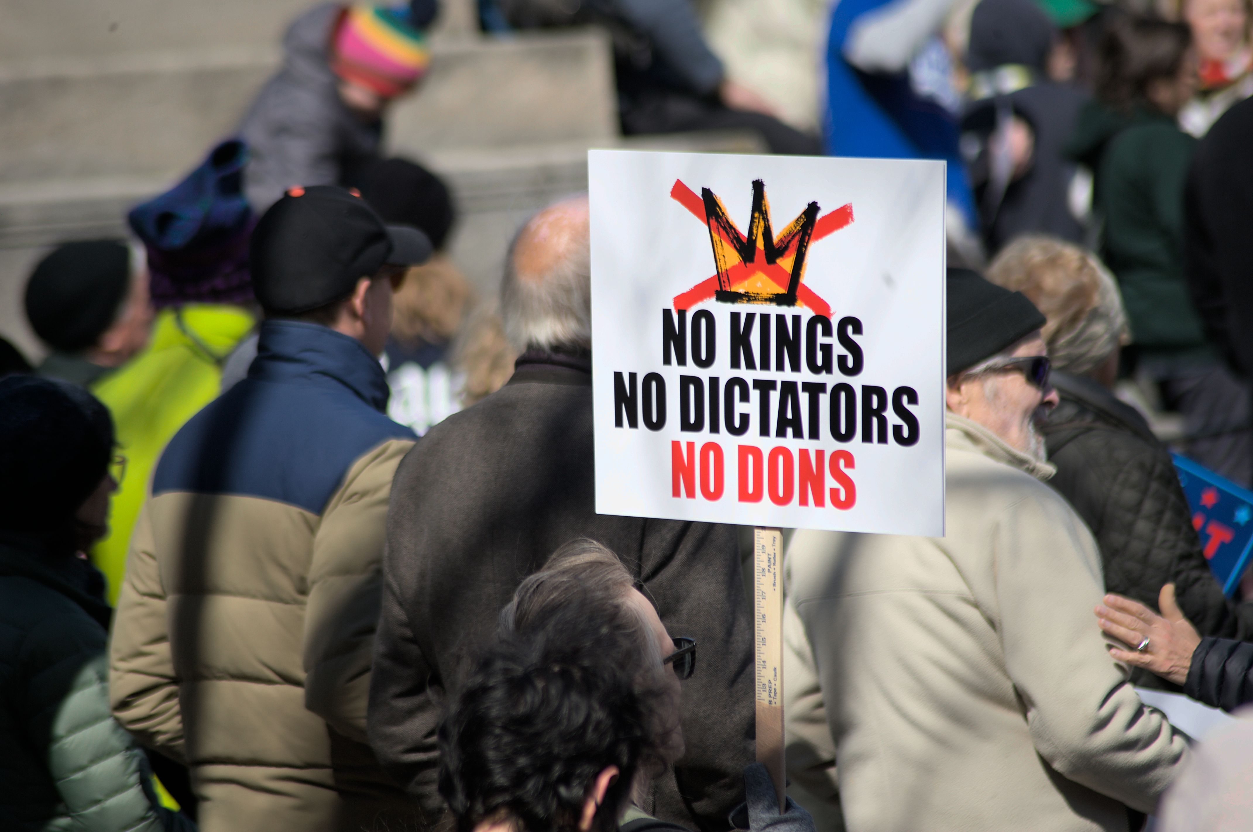 A crowd in Baltimore holds signs protesting against kings, dictators, and dons, promoting democracy.