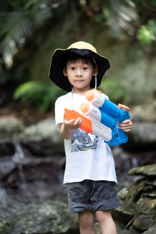 Free A young boy with a hat playing with a water gun in a natural setting, enjoying a summer day. Stock Photo