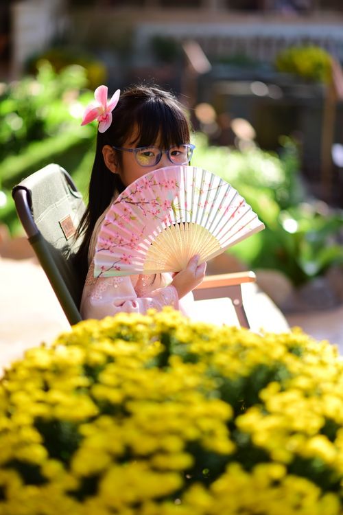 Free Young girl with glasses holding a floral fan in a sunny garden, surrounded by yellow flowers. Stock Photo
