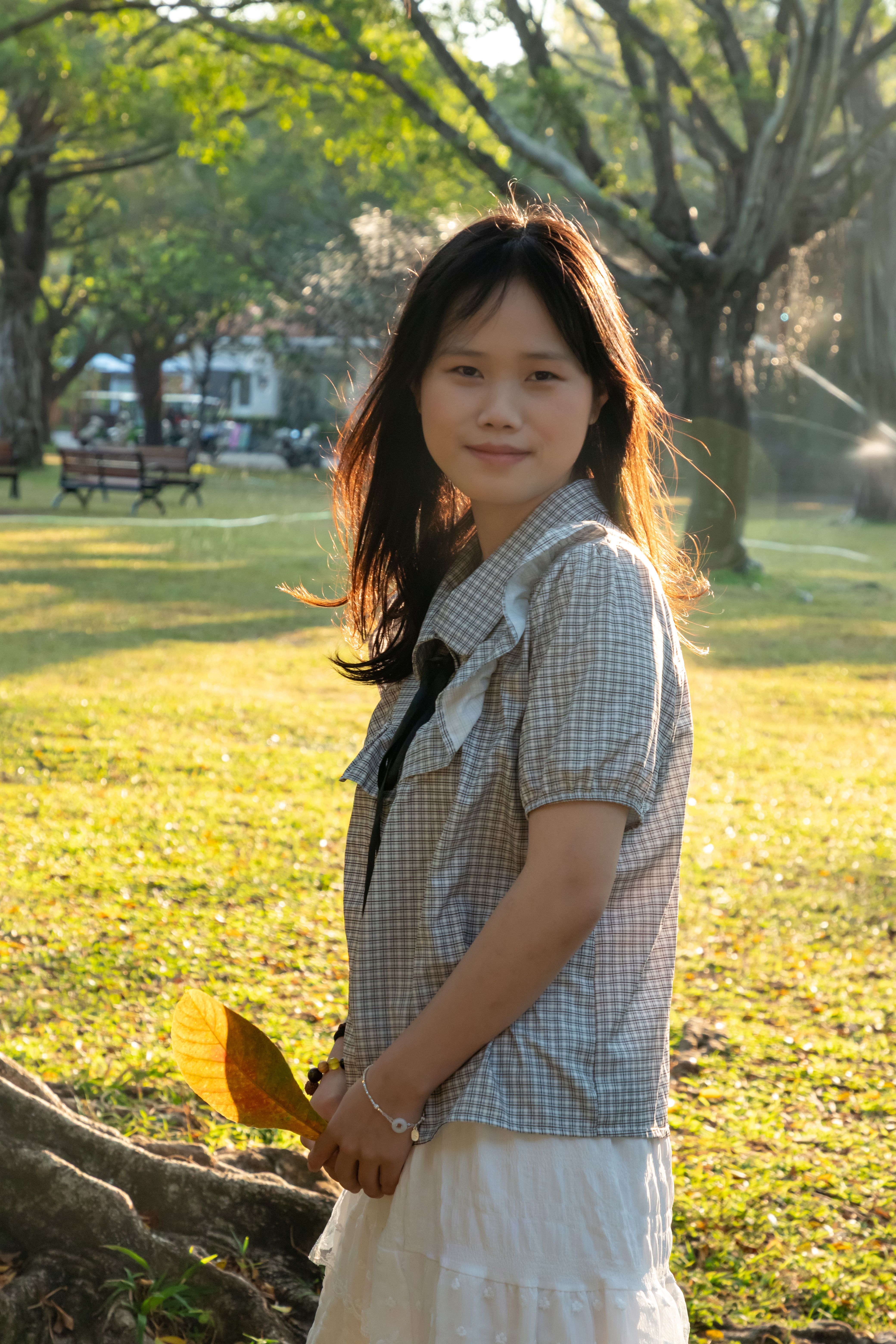 A young woman holds a leaf and enjoys the sunlight in a peaceful park setting.