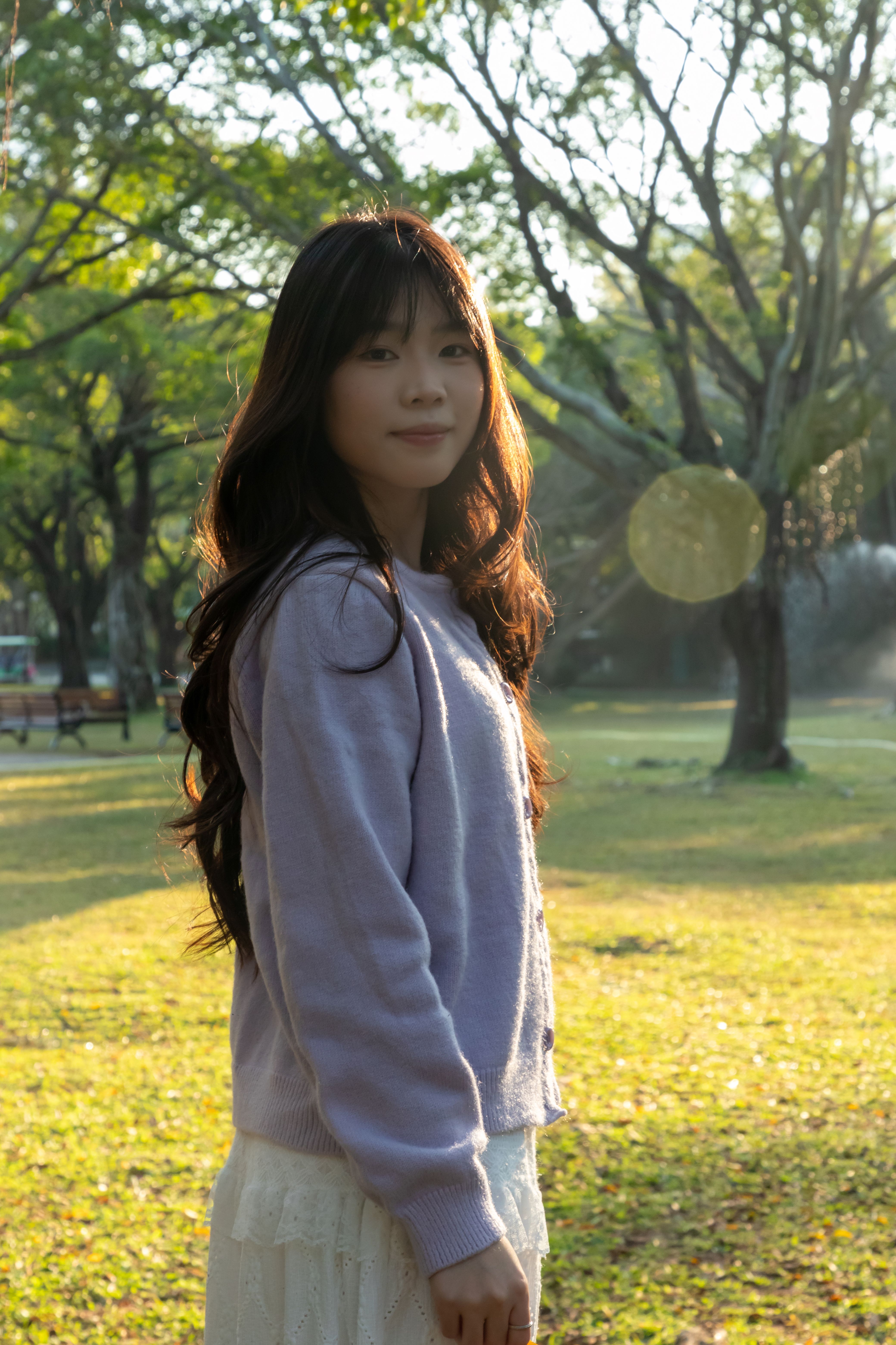 A young woman stands in a sunlit park, surrounded by lush greenery and trees.