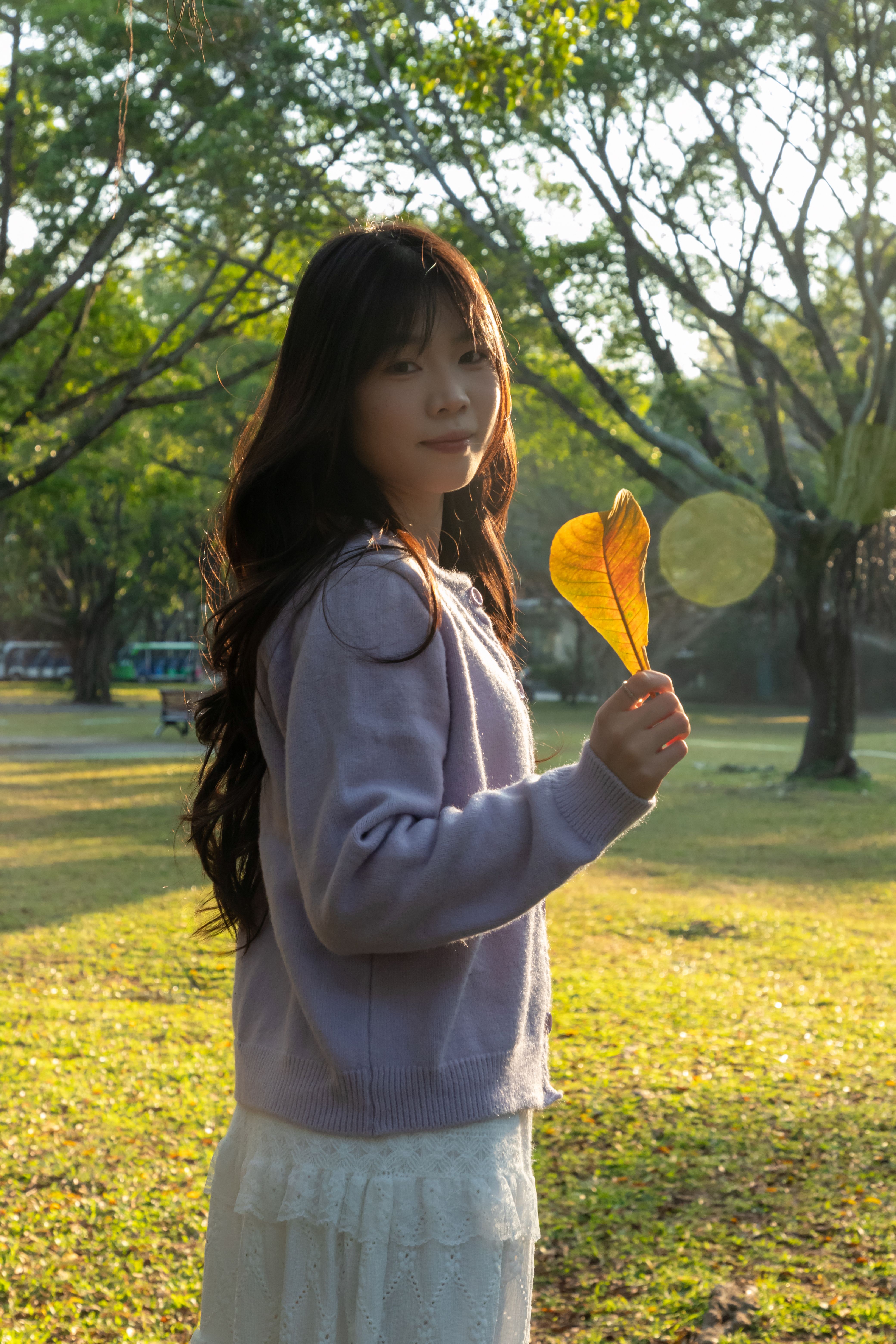 A young woman stands in a sunlit park, holding an autumn leaf, showcasing a peaceful moment.