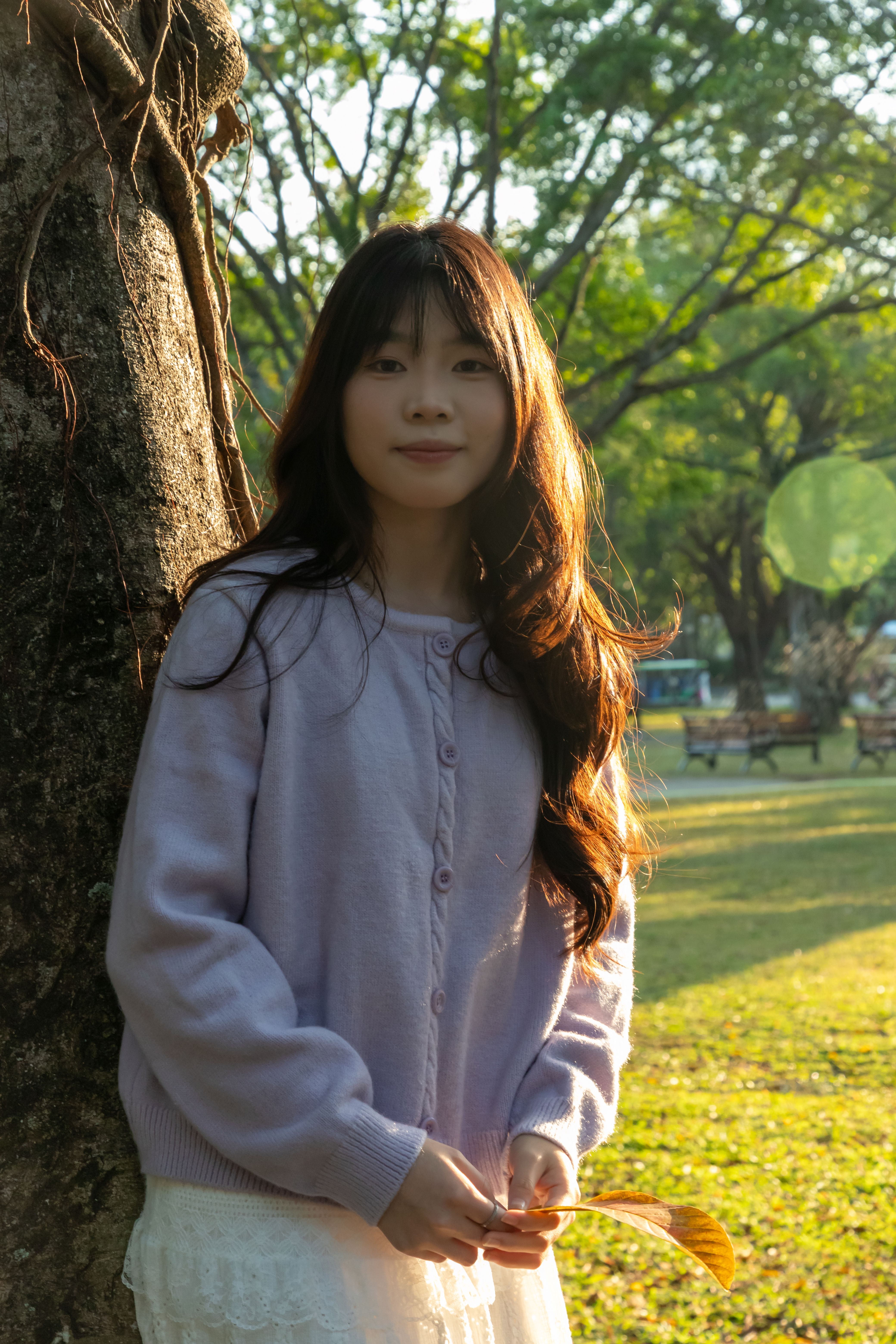 Portrait of a young woman with long hair leaning against a tree on a sunny day in a park.
