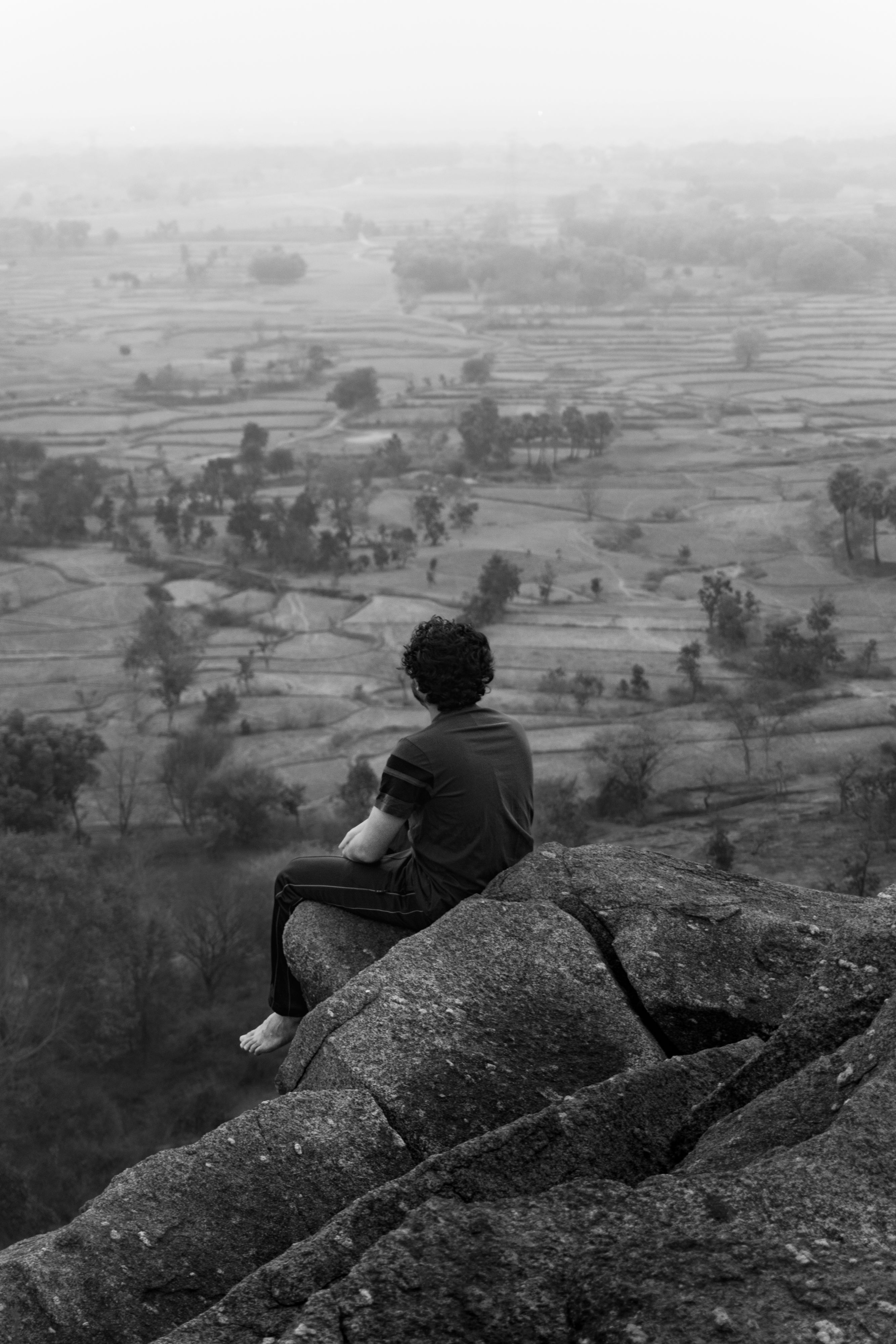 A man sits on a cliff edge overlooking a serene rural landscape, creating a sense of solitude and reflection.