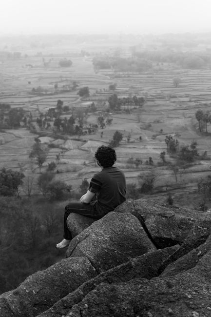 A man sits on a cliff edge overlooking a serene rural landscape, creating a sense of solitude and reflection.