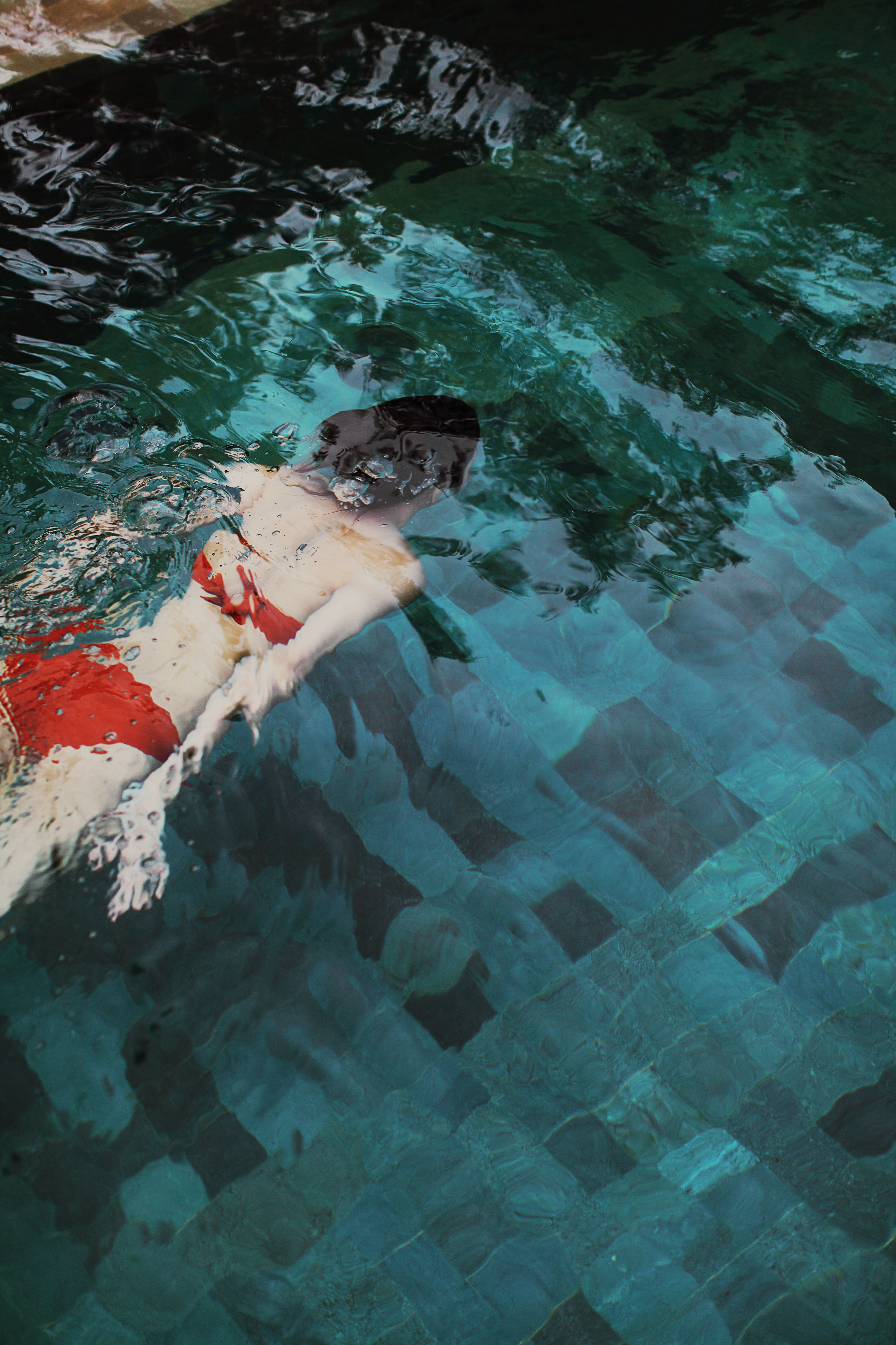Aerial view of a woman swimming underwater in a clear blue pool, wearing a red swimsuit.