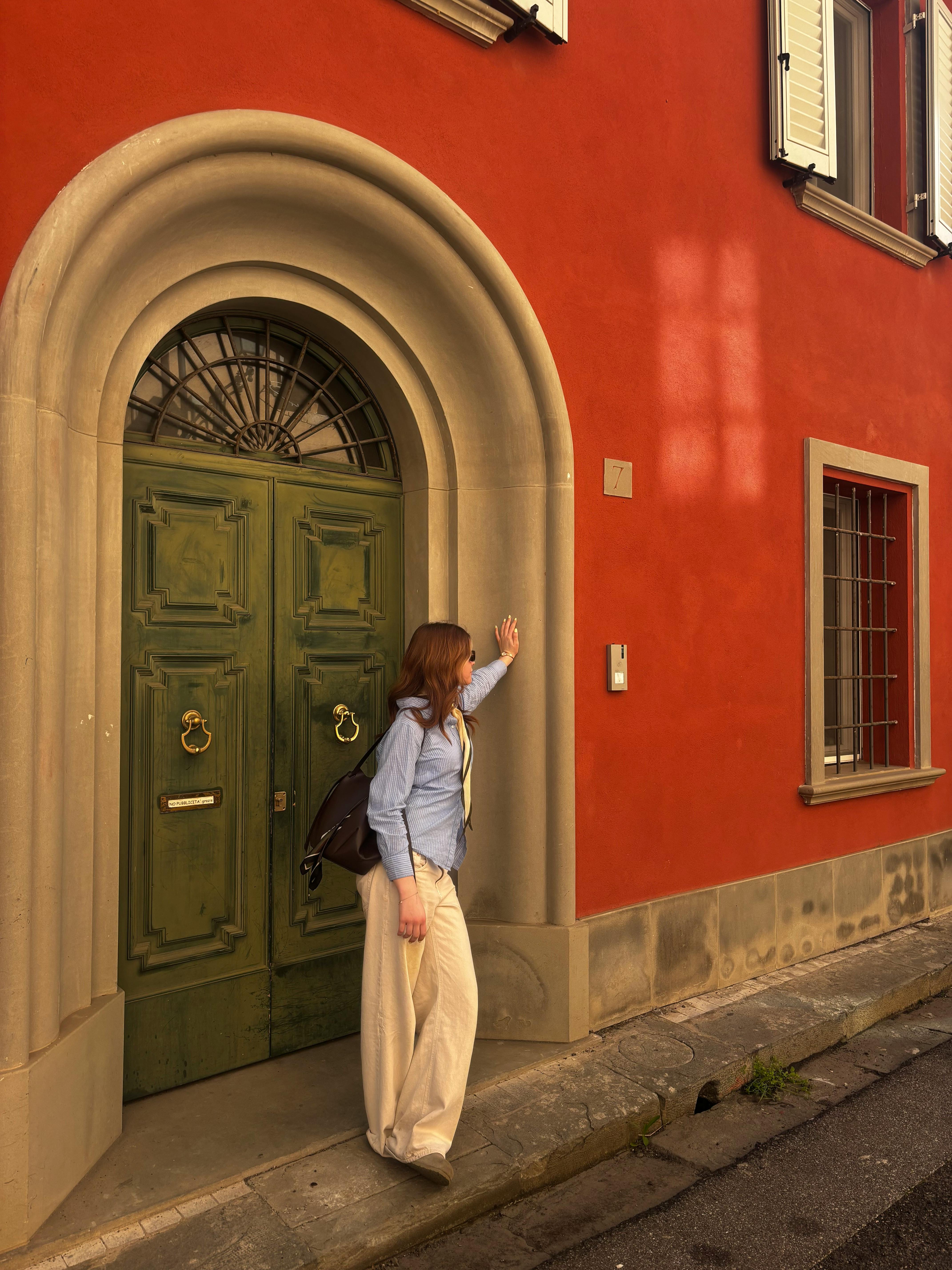 A woman in casual attire stands by a vibrant red wall with a green ornate door, creating a striking contrast.