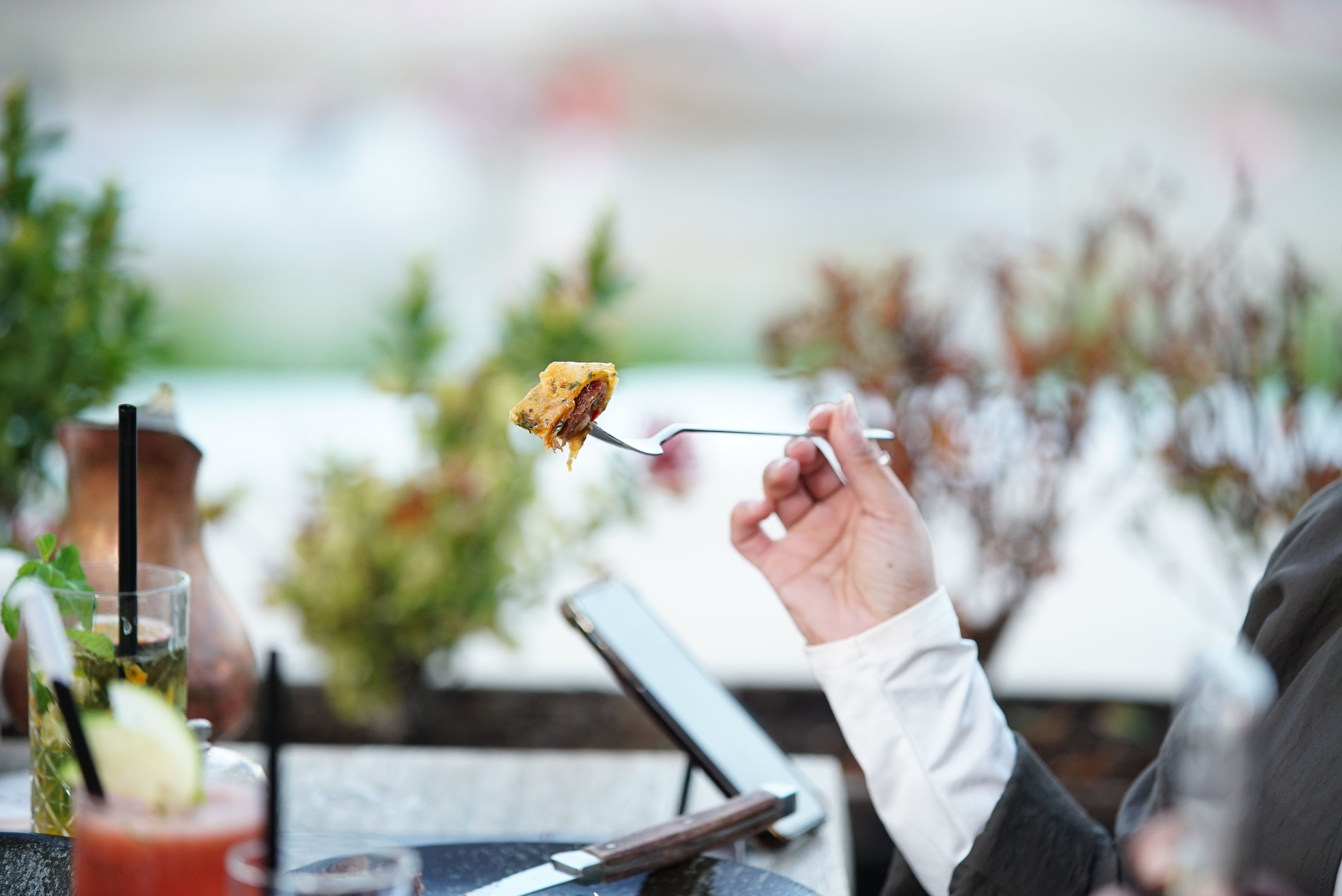 屋外で食事を楽しむ人。フォークに食べ物を乗せ、近くにはタブレット端末と飲み物がある。