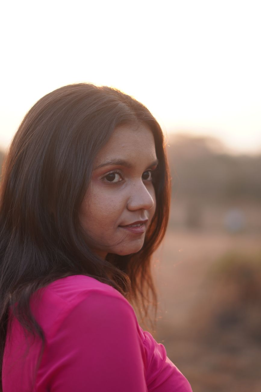 Portrait of a young woman in a pink top gazing at the camera with a serene background of a sunset outdoors.