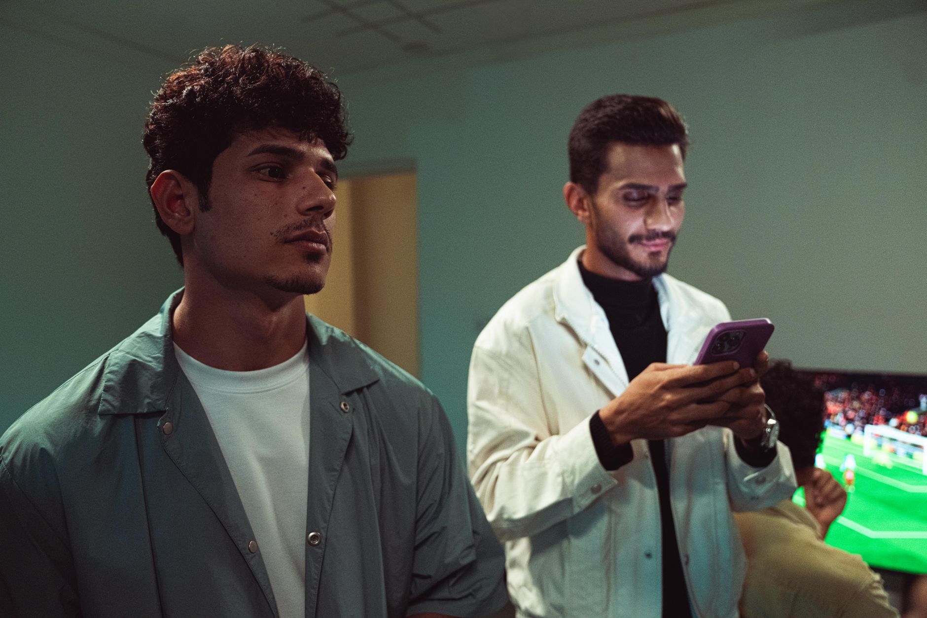 Two young men engage with smartphones and TV in a relaxed indoor setting.