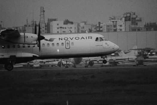 Monochrome image of a NOVOAIR ATR 72 aircraft landing at an urban airport with buildings in the background.