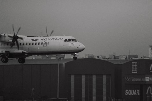 A Novoair propeller plane landing near urban structures against a twilight sky.