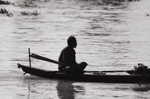Free A lone man rows a small boat on a calm river, captured in black and white. Stock Photo