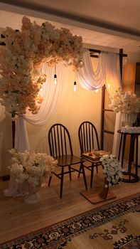Elegant floral-decorated wedding arch inside room with wooden chairs and soft lighting.