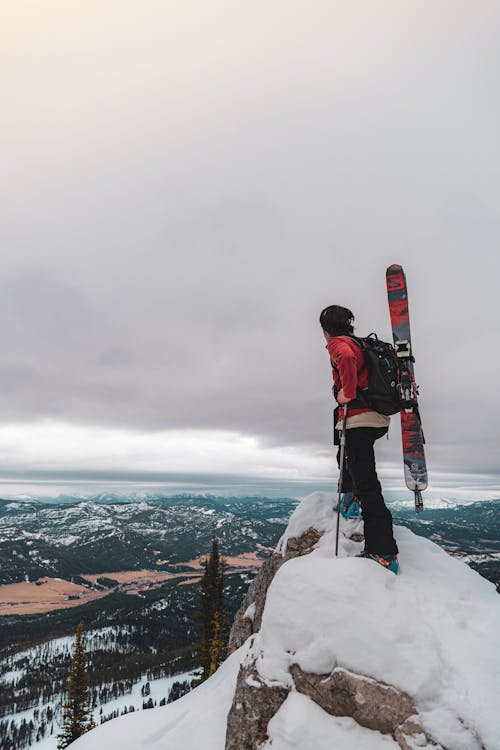 Man in Red Jacket and Black Pants Standing on Cliff with Snow