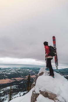 一名滑雪者站在白雪皑皑的山峰上，在阴云密布的天空下欣赏着令人惊叹的山景