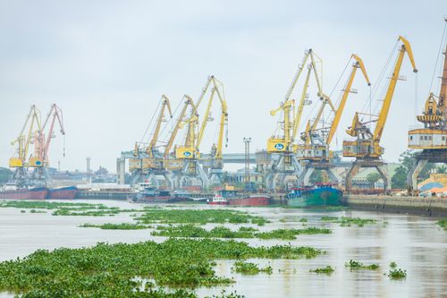 Free Large cranes at an industrial port along a river, with ships docked and greenery floating on water. Stock Photo