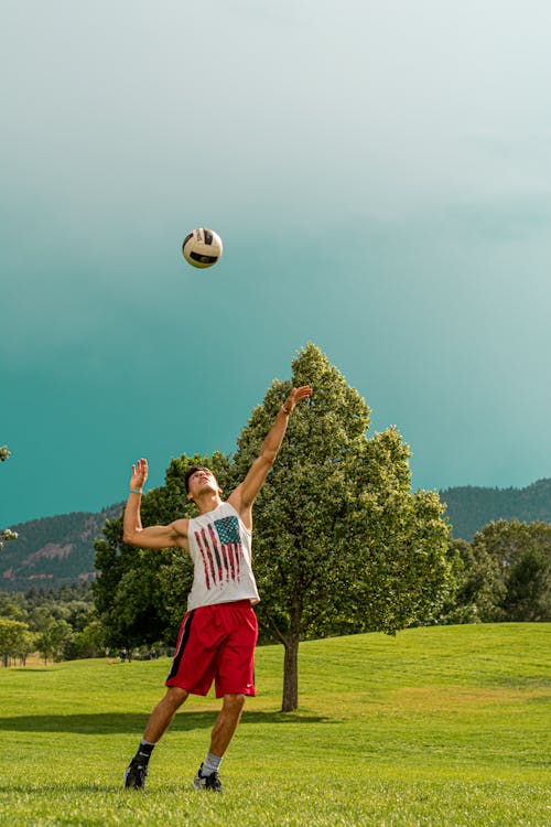 Homem De Camiseta Branca E Short Azul Em Pé No Campo De Grama Verde, Jogando Com Uma Bola De Futebol